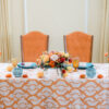 Two orange vintage highback chairs at a sweetheart table with orange patterned tablecloth, blue goblets and florals