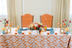Two orange vintage highback chairs at a sweetheart table with orange patterned tablecloth, blue goblets and florals