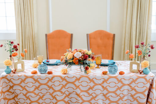 Two orange vintage highback chairs at a sweetheart table with orange patterned tablecloth, blue goblets and florals