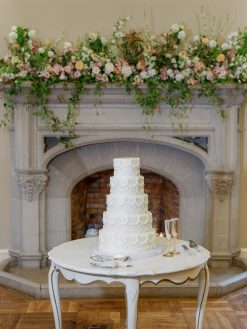 A white, ornate vintage table with curved legs holds a large tiered white wedding cake, two champagne flutes, and cake-serving utensils, set against a stone fireplace adorned with a lush floral garland.