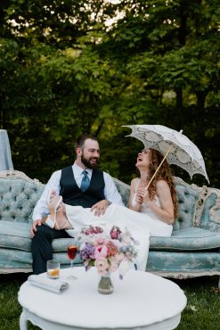 Bride and groom sitting on a vintage french light blue sofa. Bride is laughing and holding and umbrella.