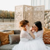 Wives in white dresses posing on a gray sofa for an outdoor photoshoot. Brown pillows. Wooden backdrop.