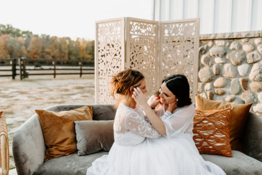 Wives in white dresses posing on a gray sofa for an outdoor photoshoot. Brown pillows. Wooden backdrop.