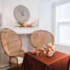 Sweetheart table at a wedding reception. Two round peacock chairs as seats. Square table with burnt orange tablecloth and florals in the center. Background is white with a window and fireplace.