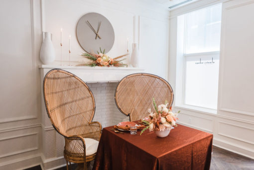 Sweetheart table at a wedding reception. Two round peacock chairs as seats. Square table with burnt orange tablecloth and florals in the center. Background is white with a window and fireplace.