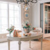 Cream colored oval vintage dining table with champagne bucket and florals. Bright window in the background.