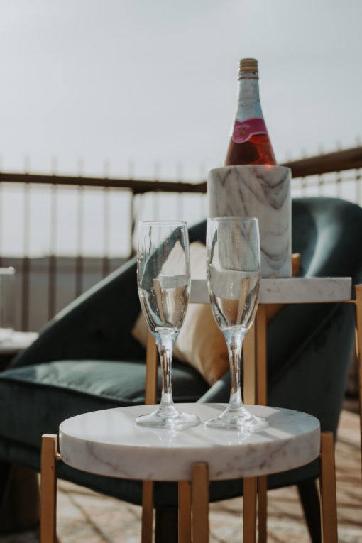 Round side table with marble top holding two champaign flutes. Behind it is a taller side table with a bottle.