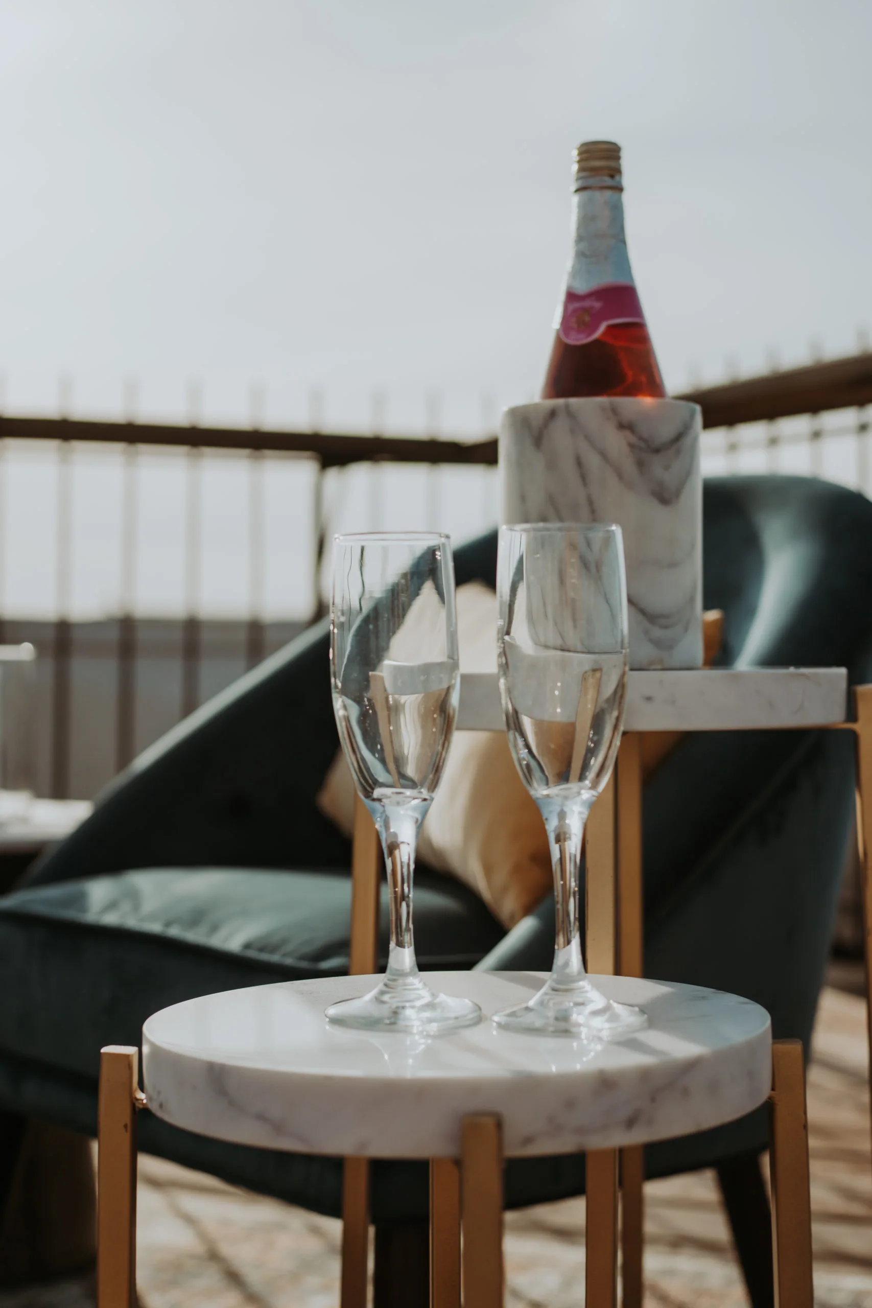 Blair petite table with champaign flutes WEBP Round side table with marble top holding two champaign flutes. Behind it is a taller side table with a bottle.