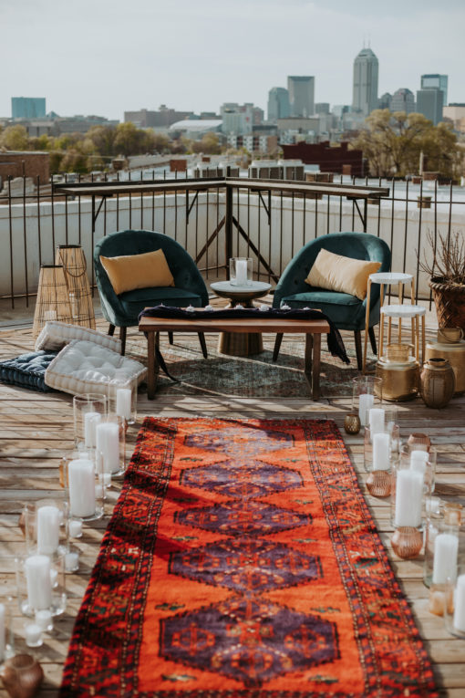 Long runner with red hues creating an aisle. Large glass cylinders with candles on the sides. Two chairs at the end with an industrial coffee table.