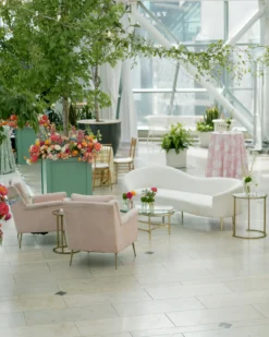 Soft seating area with white velvet sofa and two pink accent chairs. Greenery and windows in the background