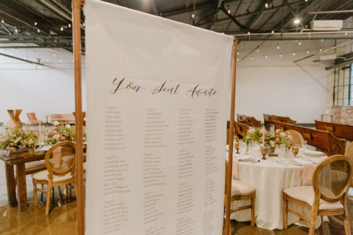 Copper rectangular sign holder with a white banner with table assignments on it in the foreground. Background is a wedding reception.