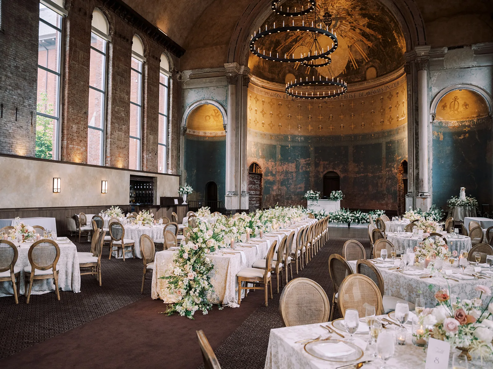 Darian and Lubo Wedding WEBP High end dining chairs with cushions in a wedding reception. One long table down the middle with round tables and square tables around.