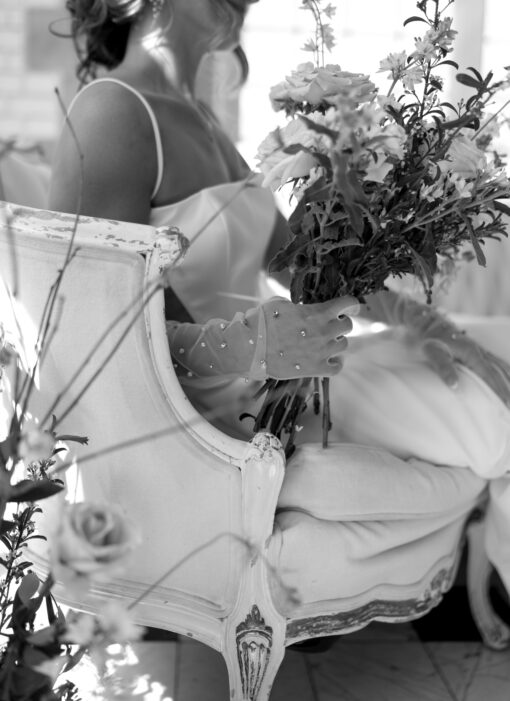 Black and white photo of a bride sitting on a vintage sofa. Wooden frame and velvet side highlighted. Bride holding florals.