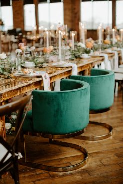 Two plush, emerald green velvet barrel chairs with golden bases are prominently featured at a wooden head table, which is elegantly decorated with flowers, candles, and place settings.