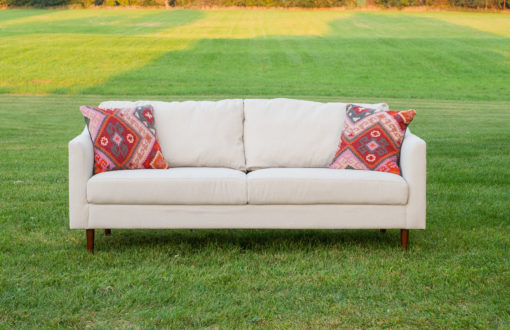 Soft off white sofa sitting on a grassy field with two multi-shades of pink pillows on each end of the sofa. Perfect setting for a photo shoot.