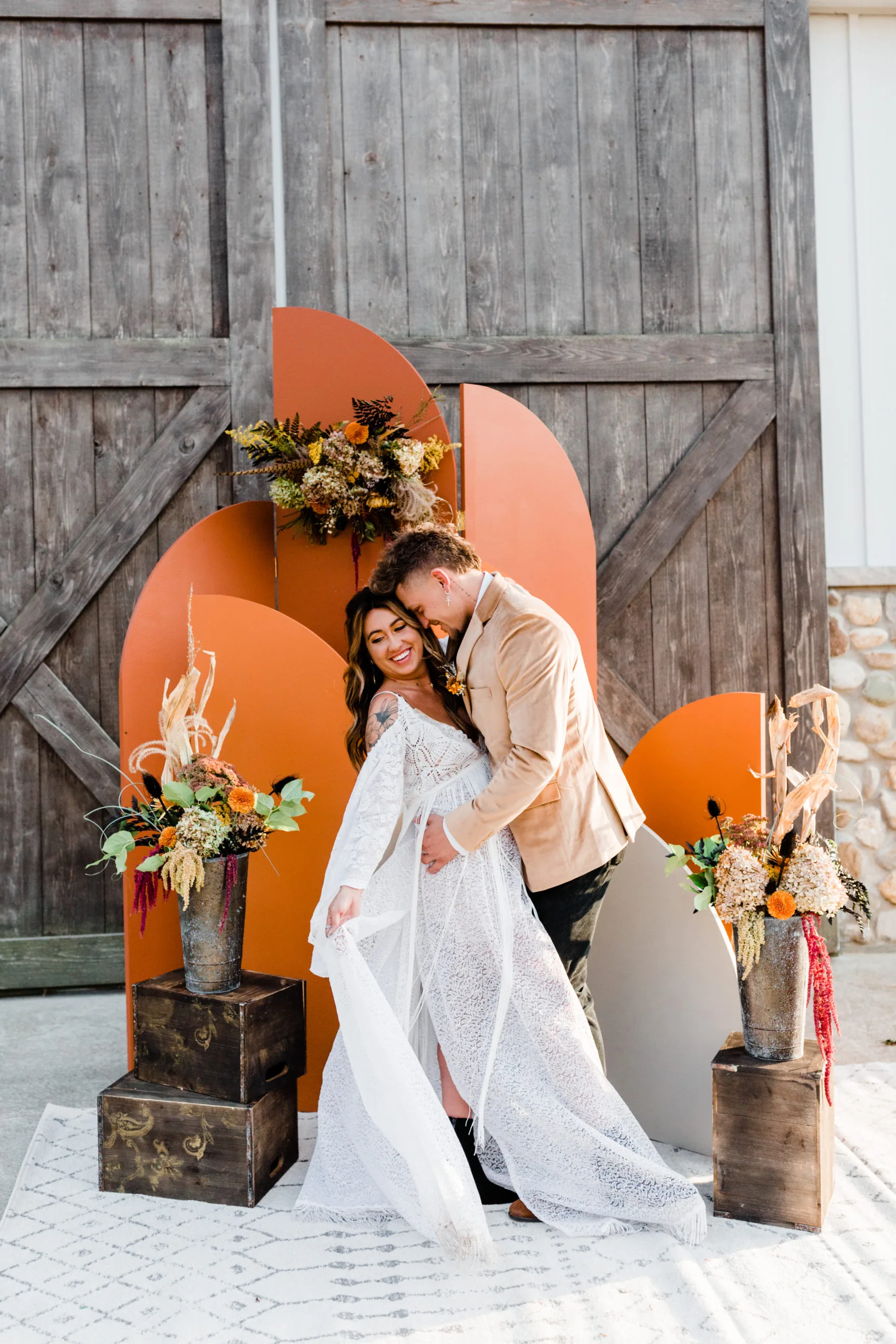 Ezra Arched Panel Backdrop and Bodrum Rug highlighting a couple in front of barn doors WEBP Couple posing for wedding photos in front of orange colored arched backdrop