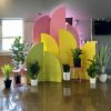 Ezra backdrop used inside for a corporate launch. Tallest set of arches is pink. Middle set is yellow. Smallest are lime green. Staggered display. Lots of plants in pots surrounding the area.