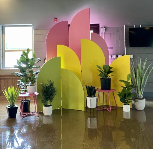 Ezra backdrop used inside for a corporate launch. Tallest set of arches is pink. Middle set is yellow. Smallest are lime green. Staggered display. Lots of plants in pots surrounding the area.