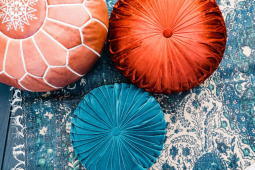 Arial close up of leather pouf, two round pillows - one teal and one rust - sitting on a teal rug.