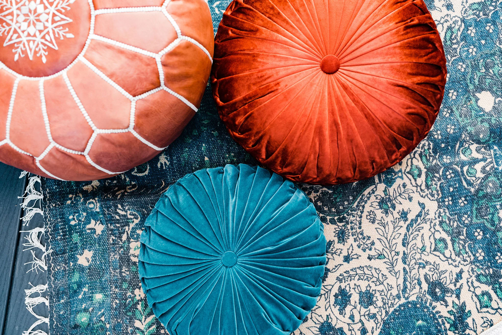 Finchley Pouf and cushions and Melina Rug WEBP Arial close up of leather pouf, two round pillows - one teal and one rust - sitting on a teal rug.