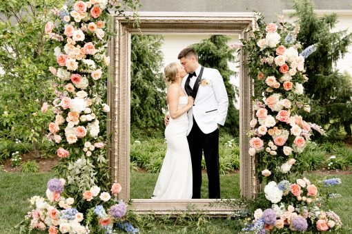 An 8 foot tall giant gold ornate frame with a bride and groom standing inside. Florals on the left and right. Outside on grass.