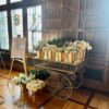 Ornate large gold metal cart with frame holding multiple vases with white flowers. Sitting on wooden floor with wooden panel walls and windows behind.