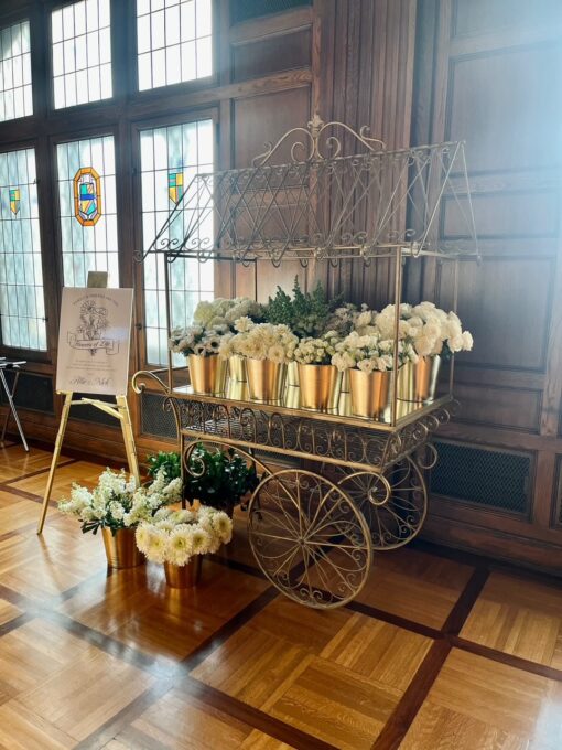 Ornate large gold metal cart with frame holding multiple vases with white flowers. Sitting on wooden floor with wooden panel walls and windows behind.