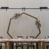 Headtable backdrop at wedding with a geometric wooden arch, wooden farm tables, and vineyard chairs against white barn doors.