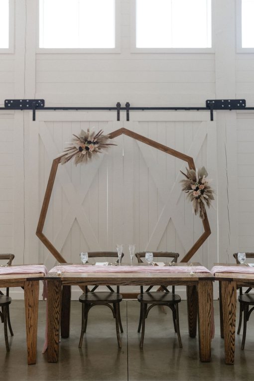 Headtable backdrop at wedding with a geometric wooden arch, wooden farm tables, and vineyard chairs against white barn doors.