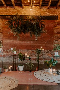 A modern event vignette with a brick wall, layered rugs, potted plants, and contemporary lounge furniture, creating an inviting atmosphere.