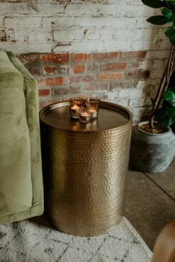 cylindrical gold drum metal side table with 3 glowing votives on top next to a green sofa against a brick wall