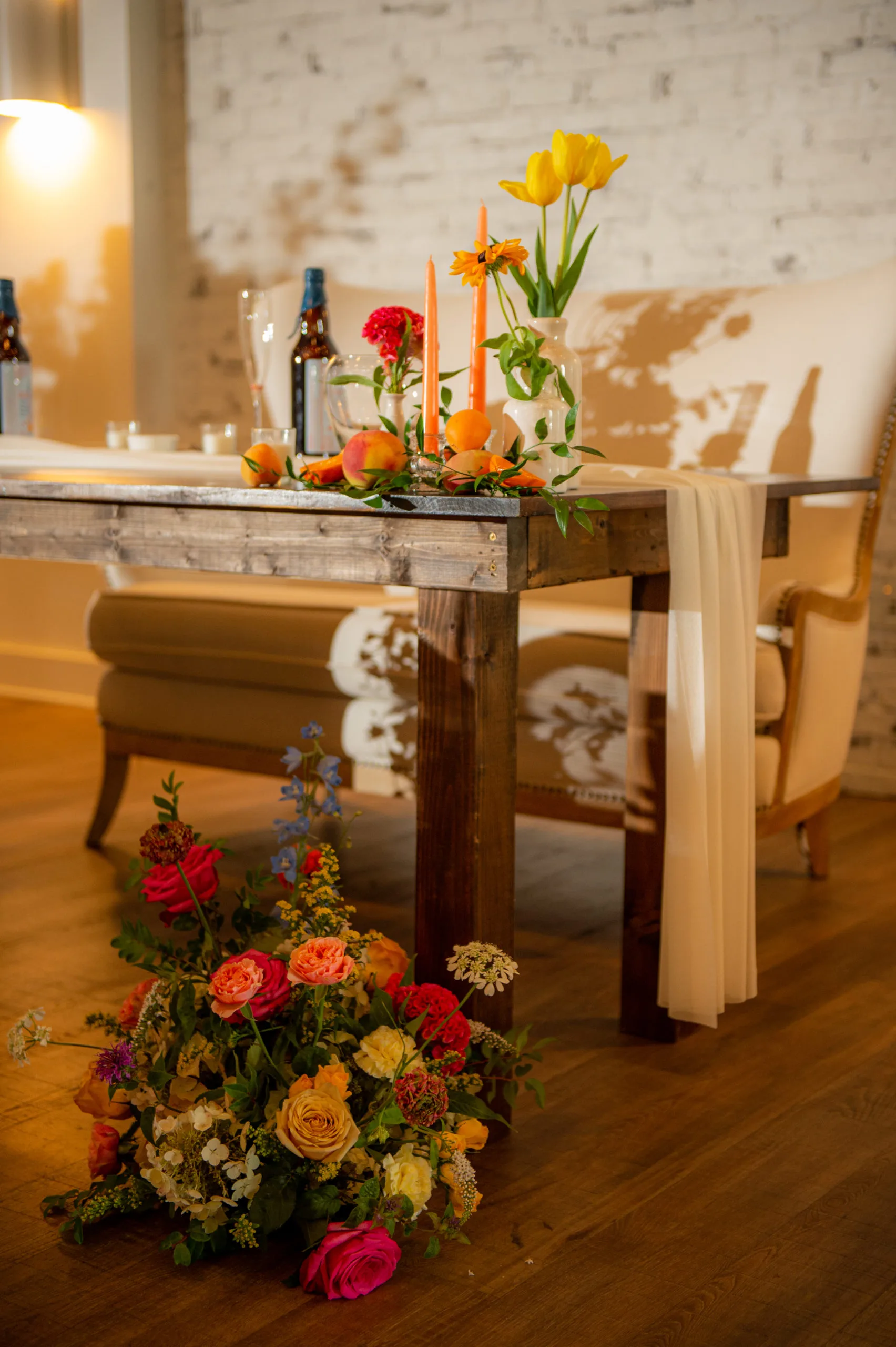 Kingston wooden farm table as a sweetheart table at wedding reception WEBP Side view of our wooden farm table used as a sweetheart table in a wedding reception. Florals at the feet and on the top. Loveseat in the back.