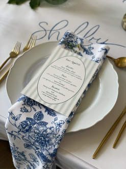 White plate with scalloped edges, blue and white napkin with a menu placed on top. Gold flatware.