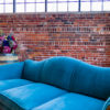 Lounge seating area featuring a vintage blue velvet sofa with wavy back and carved wooden edges. Vintage barrel to the left with florals. All against a brick wall with natural lighting.