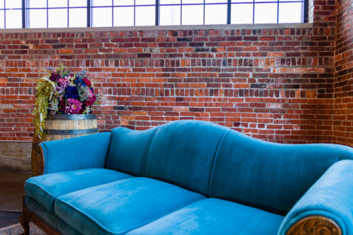 Lounge seating area featuring a vintage blue velvet sofa with wavy back and carved wooden edges. Vintage barrel to the left with florals. All against a brick wall with natural lighting.