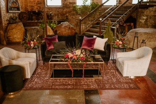 Lounge setting at a church with a black modern sofa, black two tiered glass coffee table. On the left and right, gray swivel barrel chairs. Sitting on a dark purple rug.