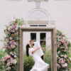 Frame in an outdoor garden with pink florals on the edges. Bride and groom kissing inside the frame. White Lilly building at Newfields in the background