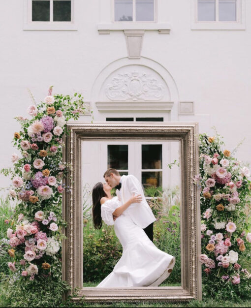 Frame in an outdoor garden with pink florals on the edges. Bride and groom kissing inside the frame. White Lilly building at Newfields in the background