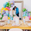 Light wood farm table in the foreground with spring tablescape and bold colors. Giant frame against the wall in the background with a bride and groom looking at each other. Colorful balloons on the edge.