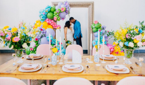 Light wood farm table in the foreground with spring tablescape and bold colors. Giant frame against the wall in the background with a bride and groom looking at each other. Colorful balloons on the edge.