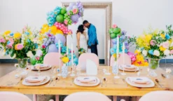 Light wood farm table in the foreground with spring tablescape and bold colors. Giant frame against the wall in the background with a bride and groom looking at each other. Colorful balloons on the edge.