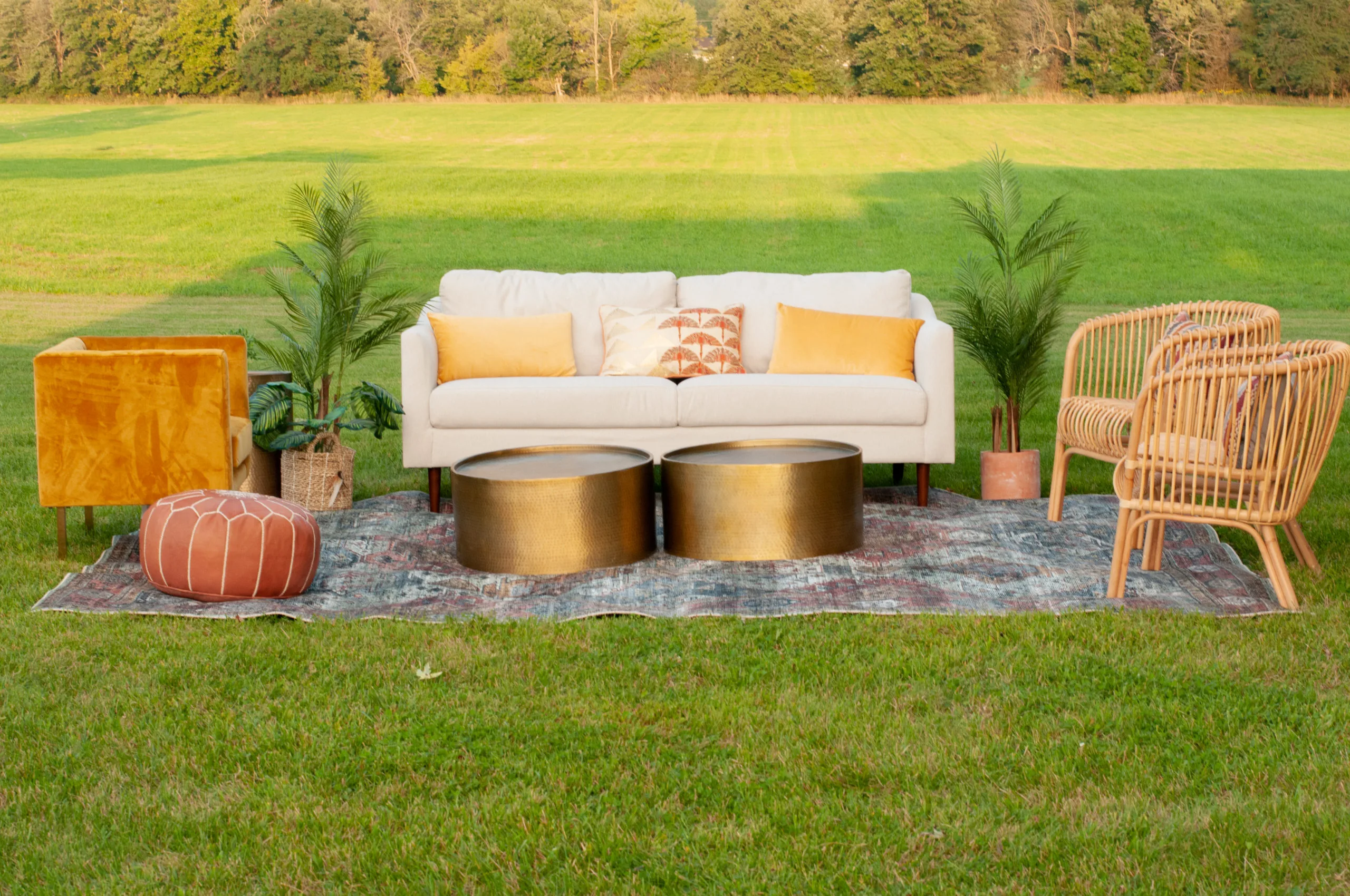 Outdoor Lounge seating with Ella Sofa, Ganzi Coffee Tables, Safiya Chairs, Finchley Pouf and Skye Rug WEBP White sofa sitting outside in a field for a photo shoot. Two gold coffee tables sitting on a rug. Two rattan chairs on the right side and a brown pouf on the left.