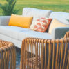 Outdoor lounge seating area on grass. Off white sofa with yellow and pink pillows. Looking over the back of two rattan arm chairs with vertical backs.