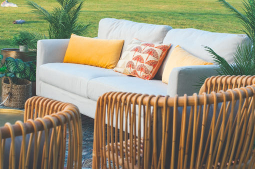 Outdoor lounge seating area on grass. Off white sofa with yellow and pink pillows. Looking over the back of two rattan arm chairs with vertical backs.