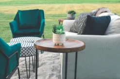 Outdoor event with styled lounge set up. Side view - looking at the side of an off white sofa with black pillow decor. Wooden topped side table with black legs. Green velvet chairs.