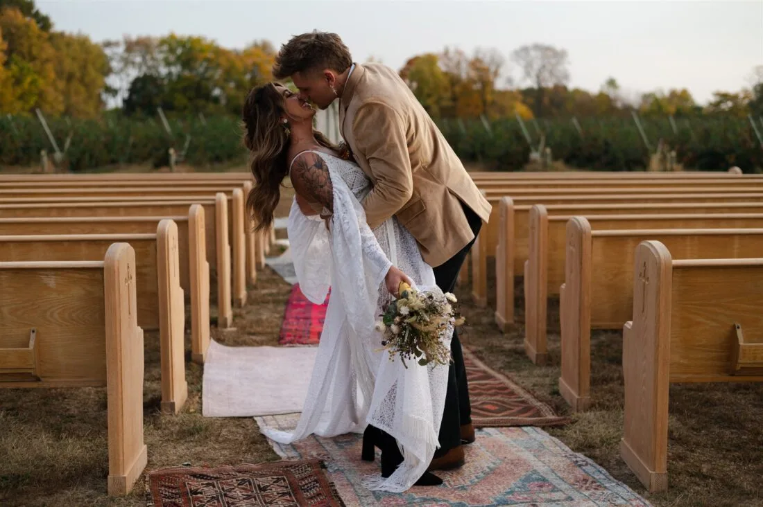 Outdoor wedding with church pews and carpet aisle - featuring Elios Rug WEBP Couple kissing in an outdoor wedding with church pews and rugs for the aisle