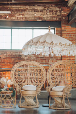 Two light rattan peacock chairs with white cushions under a boho white and gold umbrella. Brick background with a natural light window.