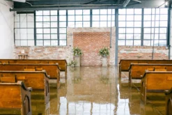 Industrial venue set up for wedding. Bank of windows up front with exposed brick wall. Pews set up for ceremony with aisle down the center.