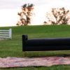 Black sofa bench with seat, arms, and no back sitting on a colorful rug in an outdoor setting. Hill in the background with a white picket fence.