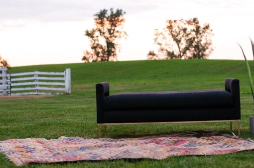 Black sofa bench with seat, arms, and no back sitting on a colorful rug in an outdoor setting. Hill in the background with a white picket fence.